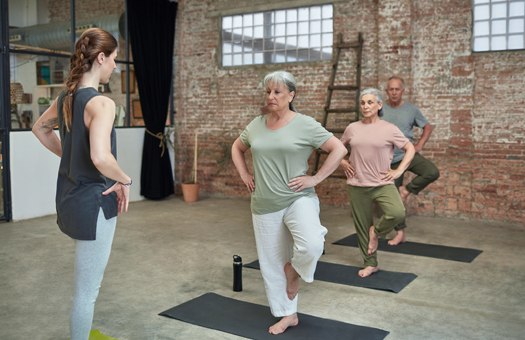 Female yoga instructor teaching senior people at health club. Active men and women are exercising on exercise mats at gym. They are in sports clothing at studio.