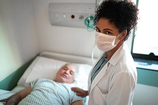 female doctor with dark curly hair wearing a mask and standing over older male patient laying in a hospital bed