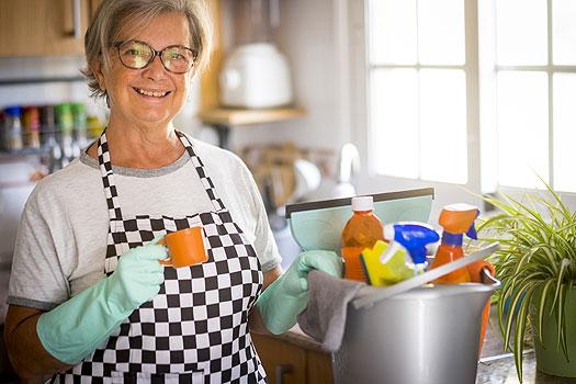 Older blonde woman wearing glasses a white tshirt a black and white checkered apron and blue rubber gloves standing in kitchen with a bucket of cleaning supplies