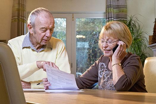 senior man and woman sitting at kitchen table reviewing paperwork and talking on a phone