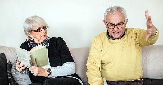 senior man and senior woman sitting on sofa, man appears to be talking expressively while woman looks up from her book